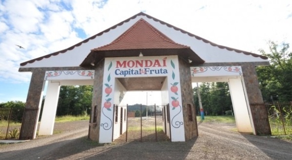 Um portal de entrada da cidade de Mondaí com o nome Mondaí Capital da Fruta escrito em vermelho e azul ao lado aparecem imagens de laranjas e a parede é branca. ao fundo tem um céu azul e algumas árvores. 