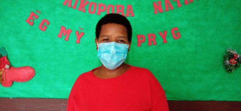 Mulher indígena negra parada em frente a um painel verde com escritas em vermelha na língua kaingang. A mulher tem o cabelo curto e usa uma camiseta vermelha e uma máscara azul. Ela está sorrindo com os olhos