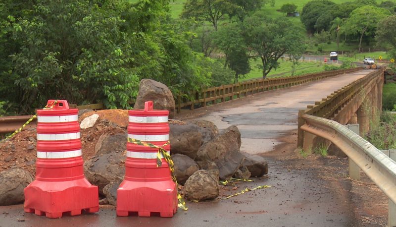 Ponte sobre o rio Chapecó fechada com pedras e terra na sua entrada e dois cones de cor laranja com listras brancos em frente e uma faixa amarelo e preto isolando o local. Do outro lado é possível ver carros parados.