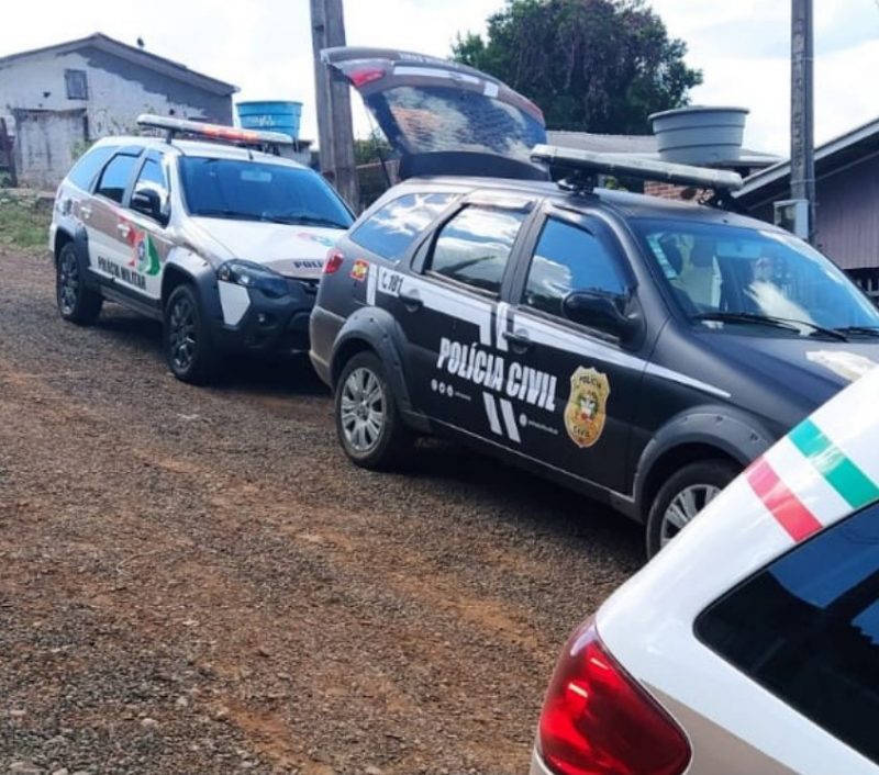 Dois carros da Polícia Militar e um carro da polícia civil com o porta malas aberto estacionados em uma rua de estrada de chão em frente a uma casa