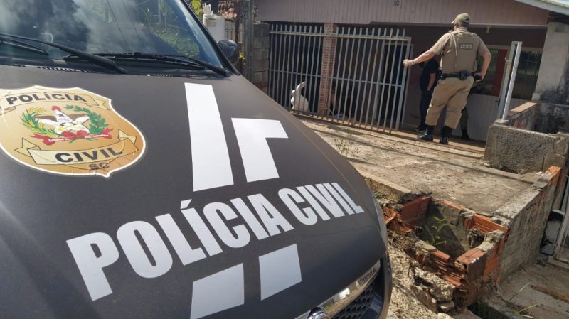 Carro da polícia civil preto com o nome em branco e o brasão estacionado em frente a uma casa, com portão cinza e casa rosa de madeira, policial parado em frente a casa,