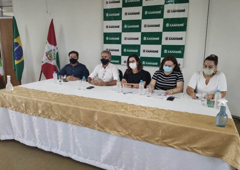 Cinco pessoas sentadas atrás de uma mesa grande. Na mesa uma toalha branca e dourada com cinco copos e cinco garrafinhas de água. Na parede um banner com o nome Xanxerê. Na mesa estão três mulheres sentadas uma ao lado da outra da direita para a esquerda. As três estão usando máscaras e com as mãos sobre a mesa. Uma delas está com a camiseta preta e a máscara branca e usando óculos, a outra está de óculos, com mascara azul e camiseta listrada em preto e branco, a terceira mulher está com o cabelo preso, de máscara verde e camiseta branca. Os dois homens sentados na outra ponta da mesa um est[a com a camisa social branca e de máscara preta, é o prefeito de Xanxerê Oscar Martarello, ele tem o cabelo grisalho. O homem ao seu lado está com uma camisa social azul escuta e uma máscara preta, seu cabelo também é preto. Todos participam da entrevista coletiva sobre a chegada das doses da vacina contra a Covid-19 em Xanxerê. Na sala também tem duas bandeiras uma do brasil e a outra de santa catarina ao lado esquerdo da foto. Em cima da mesa também tem um frasco de alcool em gel.