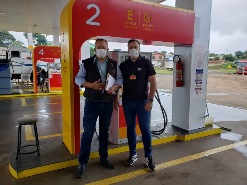Dois homens parados em frente a uma bomba de combustível em um posto de combustíveis na cidade de Xanxerê. A bomba de gasolina é vermelha com detalhes em amarelo e nela tem o número dois em branco e as letras E de etanol e G de gasolina em amarelo. Ao lado, em um palanque branco tem um extintor de incêndio vermelho e alguns cartazes colados no palanque. Do lado esquerdo da bomba tem um banco preto com a espuma de sentar cinza. Os dois homens estão de calça jeans e calçado preto. Um deles está com uma camiseta preta e um crachá amarelo e vermelho, ele usa um relógio preto no braço esquerdo e está de máscara branca, seu cabelo é preto. O outro homem, a esquerda da foto, está com uma camisa social azul claro com a manda dobrada na metade do braço e com um colete preto por cima. Ele está de máscara branca e com uma prancheta com papéis na mão esquerda. 