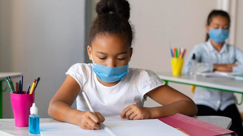 Criança sentada em frente a uma mesa na escola. Ela está com o cabelo preso em um coque e usa uma máscara azul e uma camiseta branca. Na mão ela segura uma caneta e escreve em um caderno. Ao seu lado esquerdo tem um pote rosa de canetas e lápis e ao lado um pote de alcool gel. Ao fundo aparece outra criança sentada na mesma posição olhando o caderno durante as aulas em Xaxim.