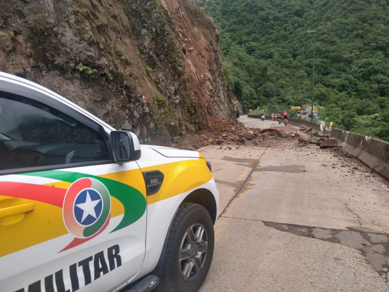 Carro da Polícia Militar branco com faixas em amarelo, verde e vermelho, com o símbolo da PM parado em frente a rodovia com rochas interditando o trânsito do local.