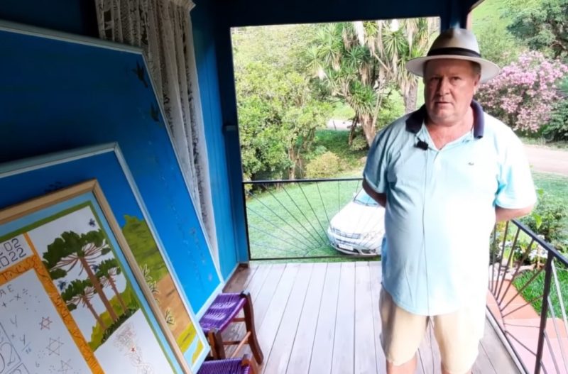 Agricultor parado ao lado direito da imagem com um calção marron e uma camisa social azul claro com as golas azul escuro e um chapéu na cabeça; ele está com as mãos para trás, ao seu lado esquerdo aparecem três quadros um atrás do outro, o maior é azul e os outros dois contém pinturas com árvores e números. Ele está na varanda de uma casa com as paredes azuis e que tem uma cortina branca na porta. Ao fundo aparecem árvores, um gramado e um carro cinza estacionado