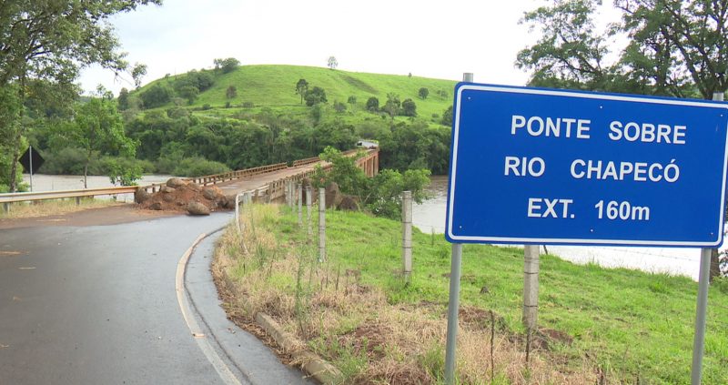 Na foto é possível ver uma parte da rodovia da integração chegando a ponte sobre o rio chapecó que está fechada com pedras e terra. Aparece também uma placa azul escrito ponte sobre o rio chapecó EXT 160 m, também aparecem árvores e um morro do outro lado da ponte