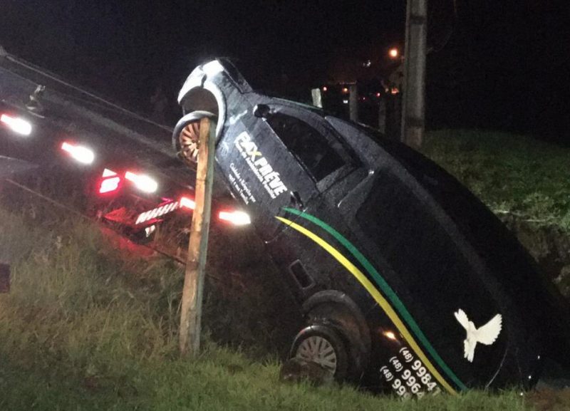 Carro funerário preto, com escritas em branco ao lado e faixas verde e amarela com o desenho de uma pomba na janela caído em um barranco de grama 