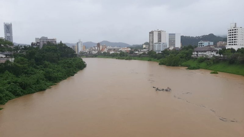 Foto do Rio Itajaí-Açu, no Centro de Blumenau. A água do está escurra, marrom, e o céu nublado. É possível observar prédios nas duas margens do rio.