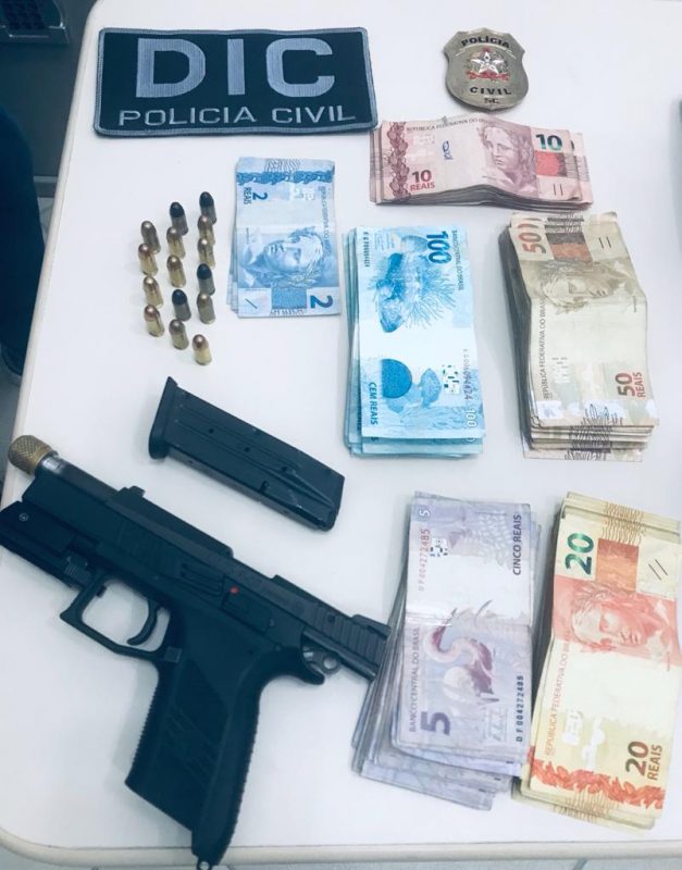Armas, munição e dinheiro em cima da mesa. Ao lado, emblema da Polícia Civil