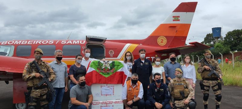 Aeronave arcanjo do corpo de bombeiros para no aeroporto de Xanxerê. A aeronave é da cor vermelho com detalhes em branco, laranja e amarelo e com a bandeira de SC, está escrito bombeiros/Samu e a porta está aberta. Na frente pessoas estão paradas entre eles o prefeito de xanxrerê e policiais, todos estão de máscara. Na frente três caixas de isopor com as vacinas contra a covid-19 e com a bandeira de SC em cima. 