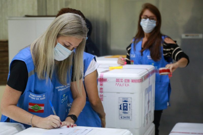 Na imagem três mulheres fazendo a separação das doses de vacina contra a Covid-19. As três estão com coletes azuis escrito secretaria de estado da saúde e com a bandeira de santa catarina abaixo nas cores vermelho, branco e verde. Elas estão usando máscaras e anotando nas caixas das doses das vacinas contra a Covid-19 que foram enviadas para Chapecó.