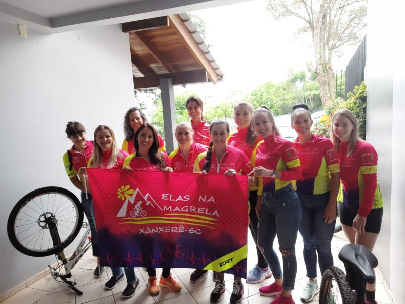 Onze mulheres vestidas com o uniforme do grupo "Elas na Magrela". Elas estão em duas fileiras, na frente cinco mulheres segurando uma bandeira escrito Elas na Magrela Xanxerê-SC e outras seis mais atrás. Todas estão posando para a foto e sorrindo. A blusa do uniforme delas é uma camiseta de manga longa cor-de-rosa com faixas amarelas nos braços e na lateral da blusa. Todas estão de tênis e algumas de calça jeans e outras de calção de andar de bike. Ao lado esquerdo e direto aparece uma parte de bicicletas.