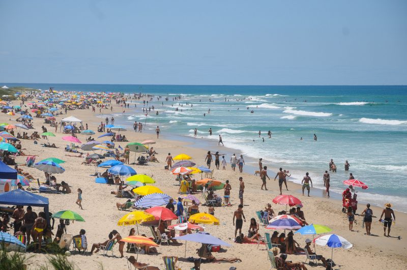 Foto da Praia do Campeche. Do lado esquerdo ao centro da foto, aparece a extensa faixa de areia clara, repleta de pessoas e guarda-sóis coloridos. Do lado direito, o mar claro. O dia está ensolarado e o céu azul. 