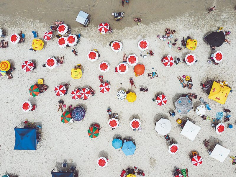 Foto aérea da faixa de areia branca da Praia do Campeche, repleta de guarda-sóis coloridos. 