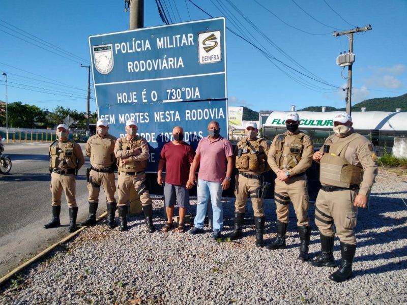 Policiais rodoviários em frente à placa que mostra o número de mortes no trecho – Foto: Divulgação/ND