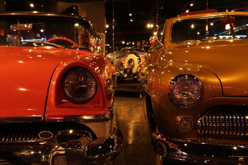 Foto do ambiente interno do Classic Car Show, Museu do Carro de Balneário Camboriú. Na foto, aparece a parte frontal de dois carros antigos, o da esquerda é vermelha e o da direita mostarda. Eles estão lustrados e brilham bastante. 