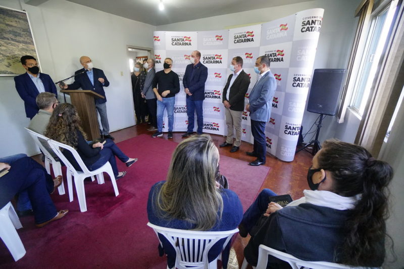 Imagem mostra pessoas em uma sala com tapete vermelho e banner do Governo de Santa Catarina