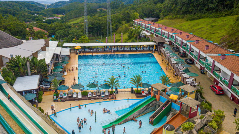 Foto aérea do Parque Aquático Cascanéia, em Gaspar (SC). No centro da foto aparecem duas piscinas. Ao redor delas há espreguiçadeiras e guarda-sóis. Em uma delas há toboáguas, que aparecem também no canto esquerdo e inferior da foto. Ao fundo aparecem morros verdes. 