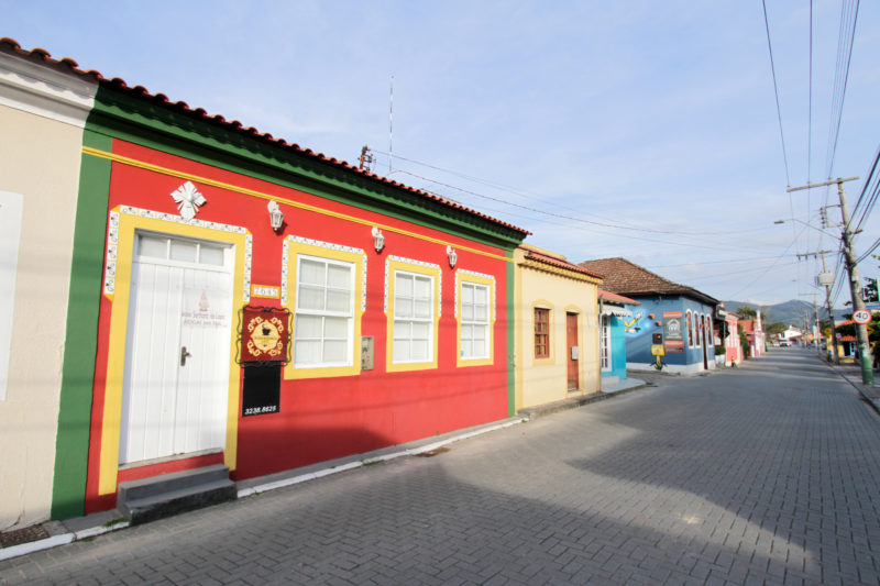 Foto da rua principal do bairro histórico de Ribeirão da Ilha, em Florianópolis. Na foto, aparece uma avenida de chão de lajotas e casas coloridas. Ao fundo, um morro verde. 