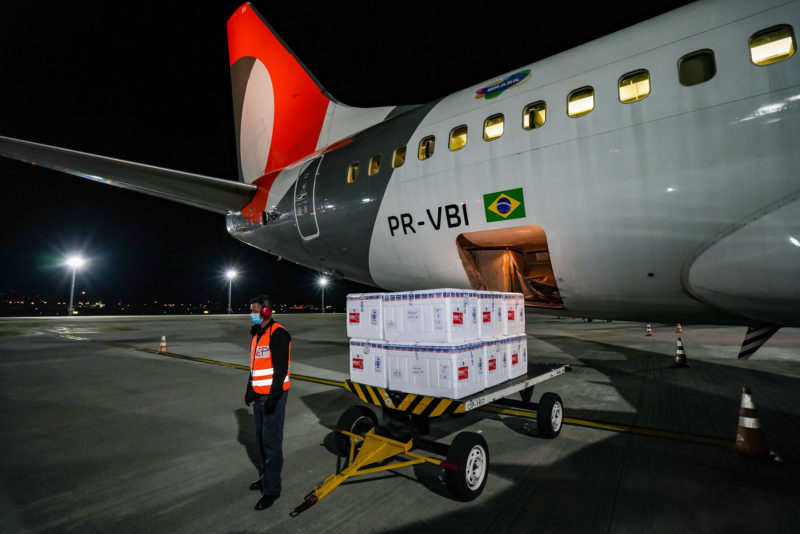 Imagem mostra caixas de isopor em carrinho e no fundo a traseira de um avião onde é possível ver uma bandeira do Brasil. Homem está próximo do carrinho com as caixas