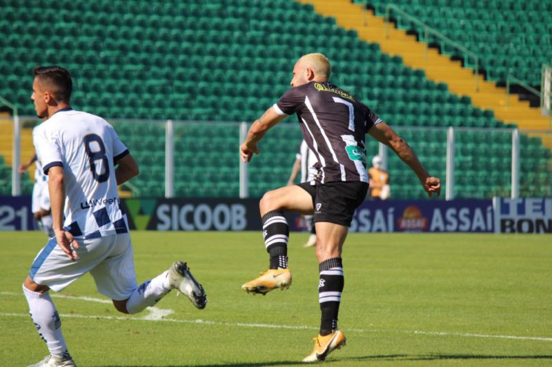 Diego Tavares após o chute e um jogador do São José-RS com a camisa branca e número oito. No fundo, placas de patrocínio onde é possível ler Sicoob