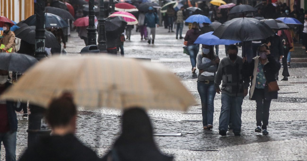 Clima chuvoso Imagem de pessoas com guarda-chuva andando em uma rua, no tempo chuvoso, para fazer alusão ao clima úmido que dará na quarta-feira