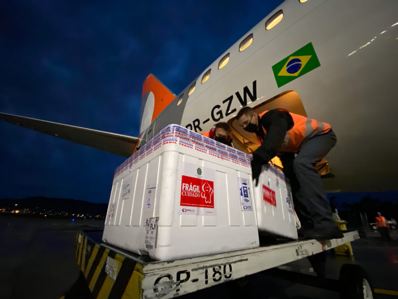 Pessoa colocando uma caixa de dose e no fundo parte do avião onde é possível ver uma bandeira do Brasil