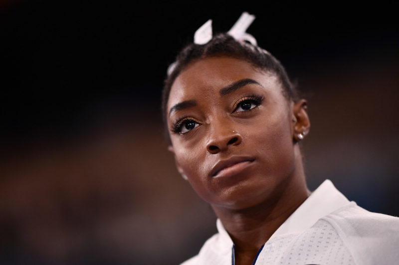 Simone Biles usa fita no cabelo e casaco branco 