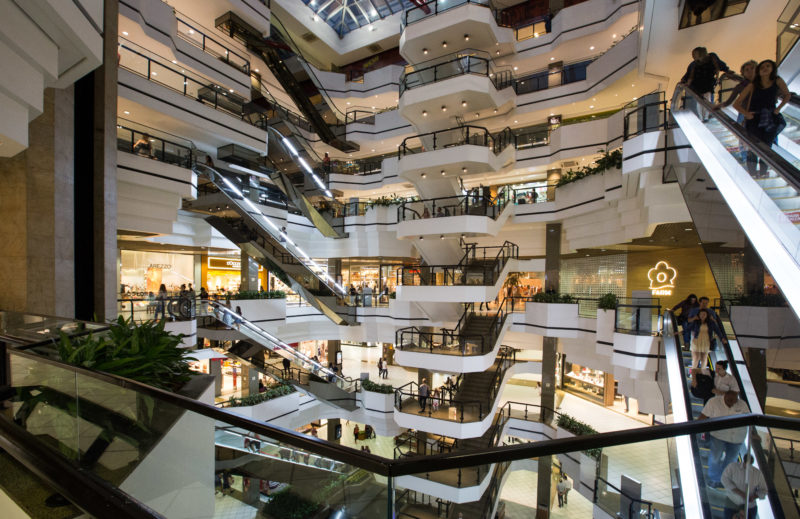 Foto interna do Beiramar Shopping em Florianópolis. No centro da foto, aparecem oito lances de escada e escandas rolantes ao redor. As paredes do prédio são brancas e os corrimãos são pretos, transmitindo uma imagem de sofisticação. Dezenas de pessoas circulam pelo shopping. 