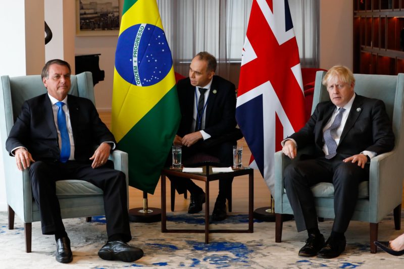 Imagem mostra Presidente Bolsonaro na esquerda, ao lado da bandeira do Brasil, no meio o traduro, ao lado a bandeira do Reino Unido e o primeiro-ministro Boris Johnson. Todos estão com terno preto