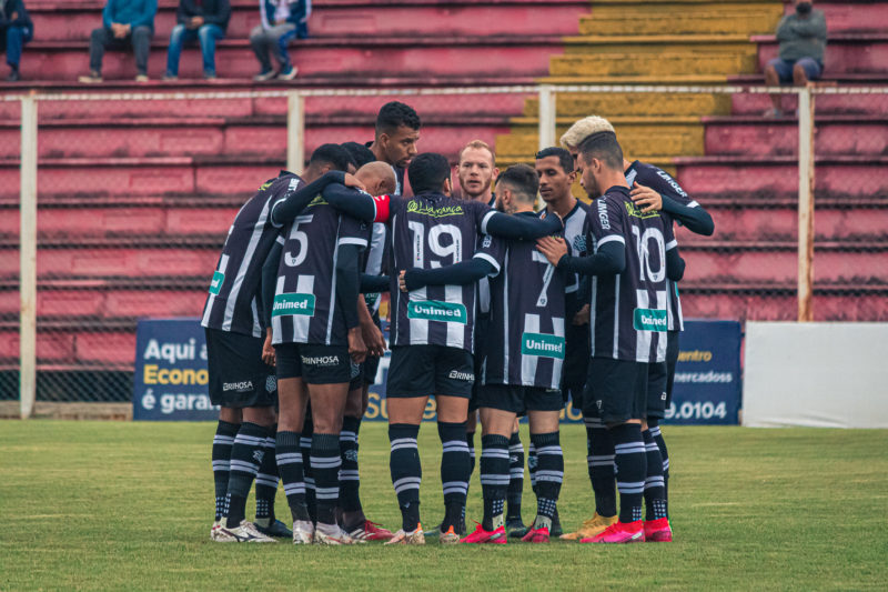 Imagem mostra jogadores do Figueirense reunidos em círculo, com camisa listrada e no fundo aparece a arquibancada em vermelha