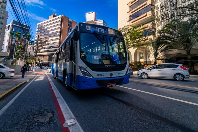 Imagem mostra ônibus de florianópolis da cor azul e branco parado e no fundo um carro branco. o céu está sem nuvens