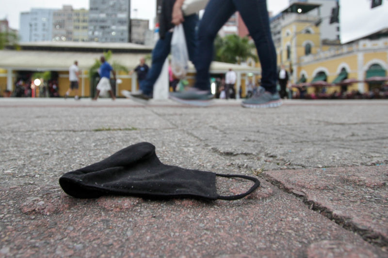 Imagem mostra máscara no chão de rua enquanto no fundo passam duas pessoas andando