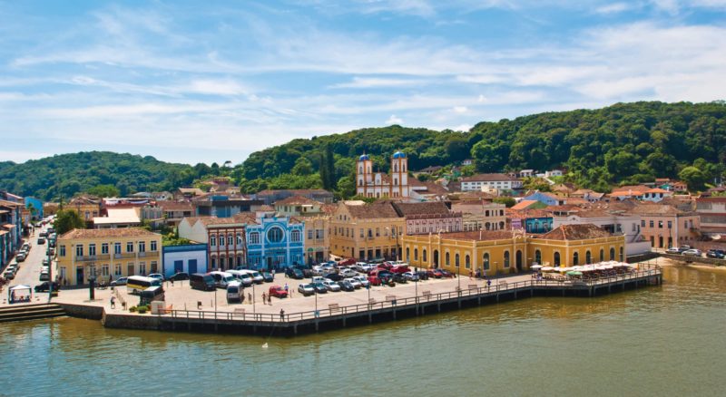 Foto aérea do Centro Histórico de São Francisco do Sul. No centro da foto, vários casarões antigos coloridos. À frente, as águas tranquilas da Baía da Babitonga e ao fundo morros verdes. 