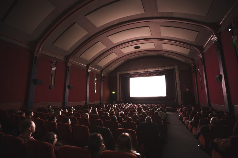 Foto mostra pessoas sentadas em poltronas em sala de cinema e tela branca