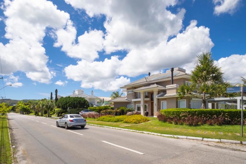 Foto de uma rua do bairro de Jurerê Internacional. Do lado direito, uma mansão com um amplo jardim. Um carro está na rua. Ao fundo, aparecem outros jardins. O céu está bem azul, com nuvens. 