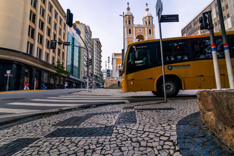 Imagem mostra calçada em primeiro plano, depois o lado de um ônibus da cor amarela e no fundo uma igreja