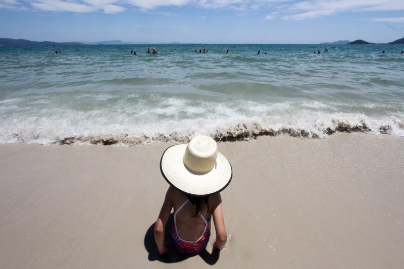 Imagem mostra criança com um chapéu próximo da água e sentada na areia