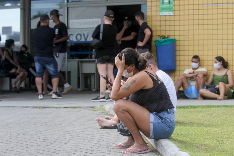 Limite diário de atendimentos em postos de saúde fazia com que pessoas esperassem em fila horas antes da unidade abrir para tentar atendimento médico, foto mostra mulher sentada no meio fio, de máscara, com mão na cabeça. Ao fundo um posto de saúde e outras pessoas esperando em pé e sentadas no chão