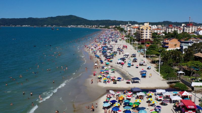 Foto da Praia de Canasvieiras, em Florianópolis (SC), no verão de 2002. Na metade à esquerda da foto aparece o mar cheio de banhistas. À direita, a faixa de areia branca com guarda-sóis. Depois dela, uma faixa de vegetação e prédios. Ao fundo, morros verdes.