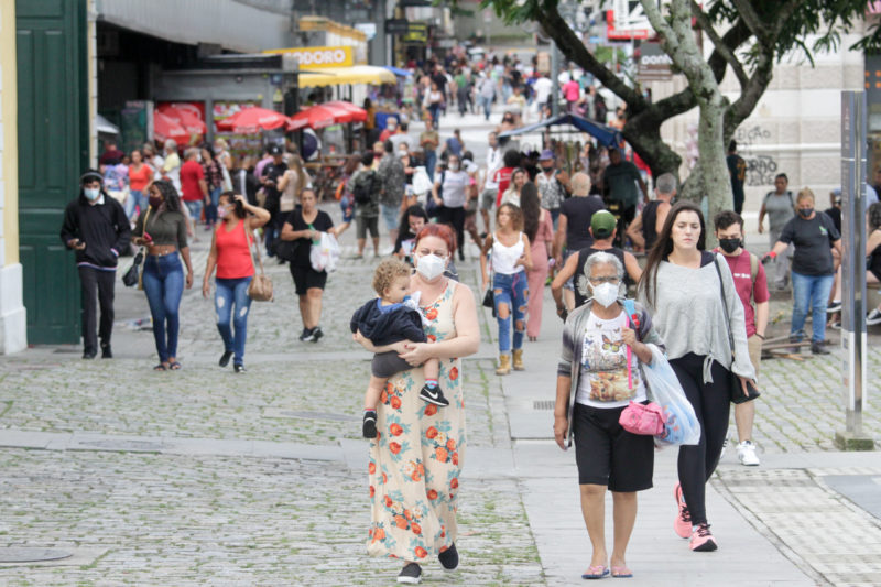 Imagem mostra pessoas em uma rua de calçamento em SC e algumas estão usando máscara. Em primeiro plano aparece uma mulher com uma criança no colo