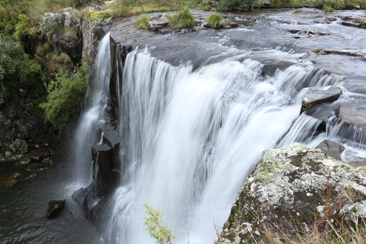 Foto de uma cachoeira pequena, larga e volumosa. A água escorre por rochas escuras até um poço. No canto esquerdo, aparece parte da vegetação.