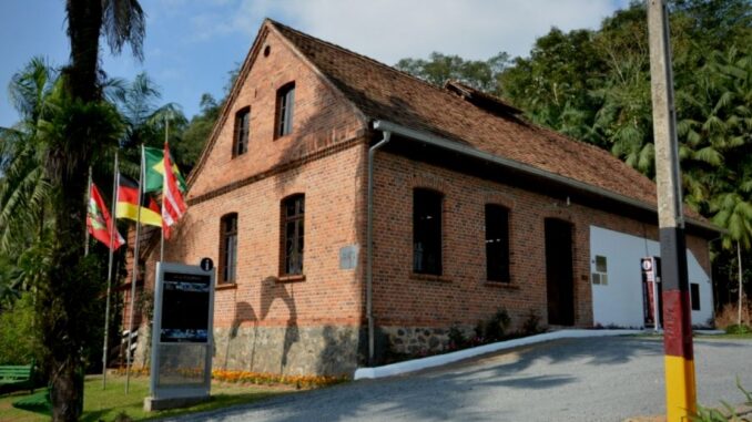 Foto da fachada do Centro Cultural da Vila Itoupava, com as bandeiras da cidade, Estado, Brasil, Alemanha hasteadas 