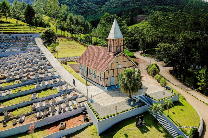 Foto de uma Igreja Luterana, na cidade de Benedito Novo, construída em estilo enxaimel, com tijolos à vista e ripas de madeira sobrepostas. Ela está no alto de uma colina. No lado esquerdo há um pequeno cemitério e no lado direito e atrás muitas árvores e morros verdes.