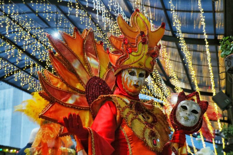 Foto de uma pessoa fantasiada e mascarada. A fantasia é em tons de laranja e amarelo e tem vários ornamentos. A pessoa segura uma outra máscara na mão esquerda e tem a mão direita esticada. 