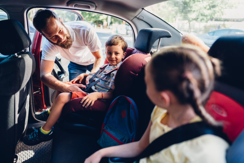 Na imagem, um homem cuida da segurança das crianças no banco de trás do carro para curtir as férias em família