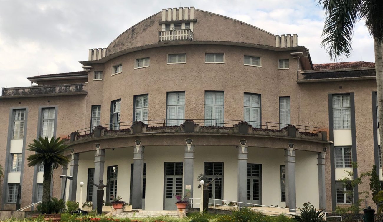 Foto horizontal mostra a fachada do Teatro Carlos Gomes. O centro da fachada é arredondado, em marrom claro e com sete janelas verticais.