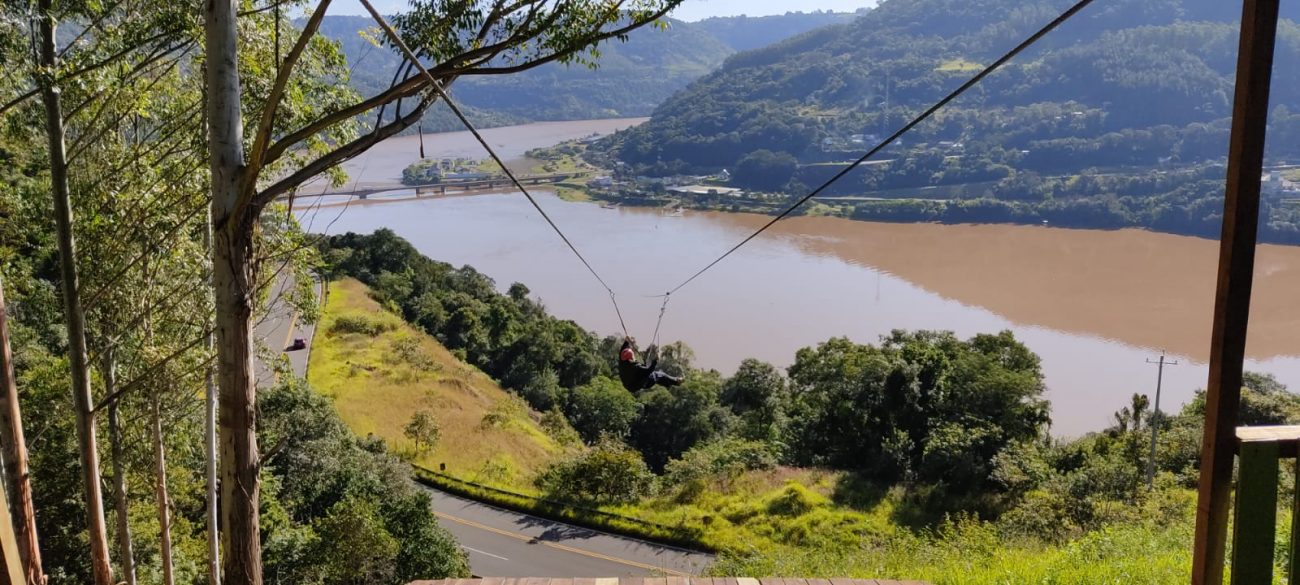 Foto da tirolesa interestadual que liga Chapecó ao Rio Grande do Sul. No centro da foto, dois cabos onde uma pessoa desce presa. Ao fundo, uma via de asfalto, o rio Uruguai e morros verdes.