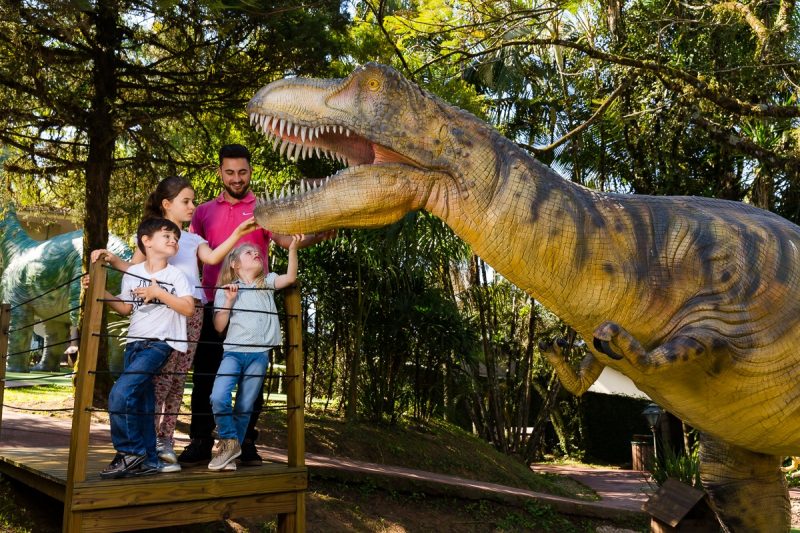 Na imagem, aparecem três crianças e um adulto interagindo com um dinossauro de brinquedos, como sugestão de diversão nas férias escolares